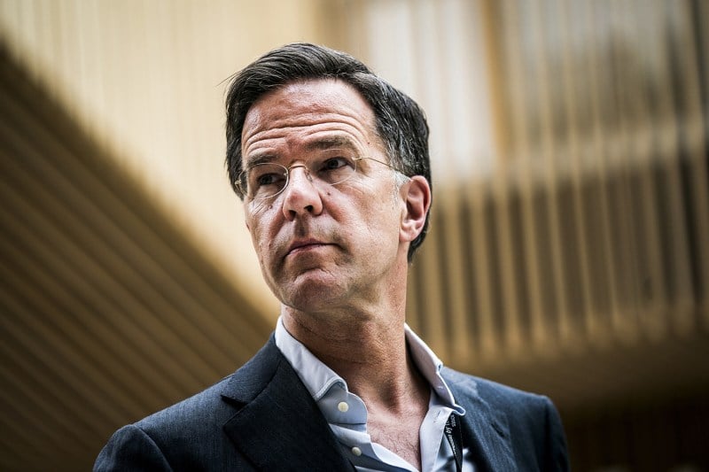 Outgoing Dutch Prime Minister Mark Rutte, wearing wire-frame glasses, a suit jacket, and open-collared button-up shirt with no tie, furrows his brow as he looks to his right.
