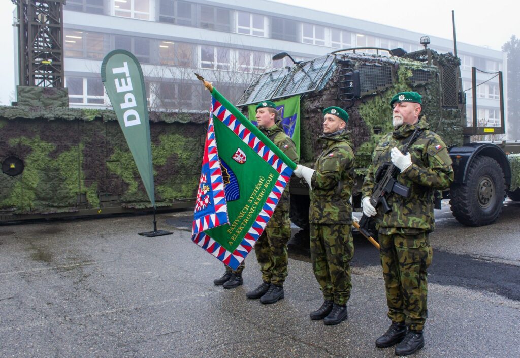 Four soldiers in camouflage stand in attention, facing left. The third from the left is holding a green flag that appears to be the standard banner of an army regiment. It