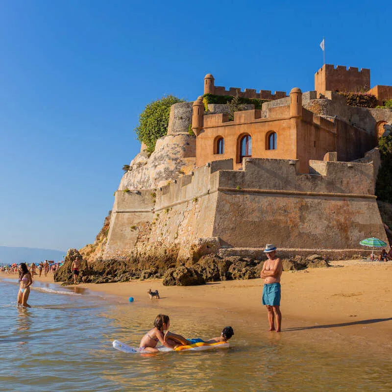 Tourists Bathing In A Sandy Beach Near A Medieval Castle In Ferragudo, Algarve, Southern Portugal, Southern Europe