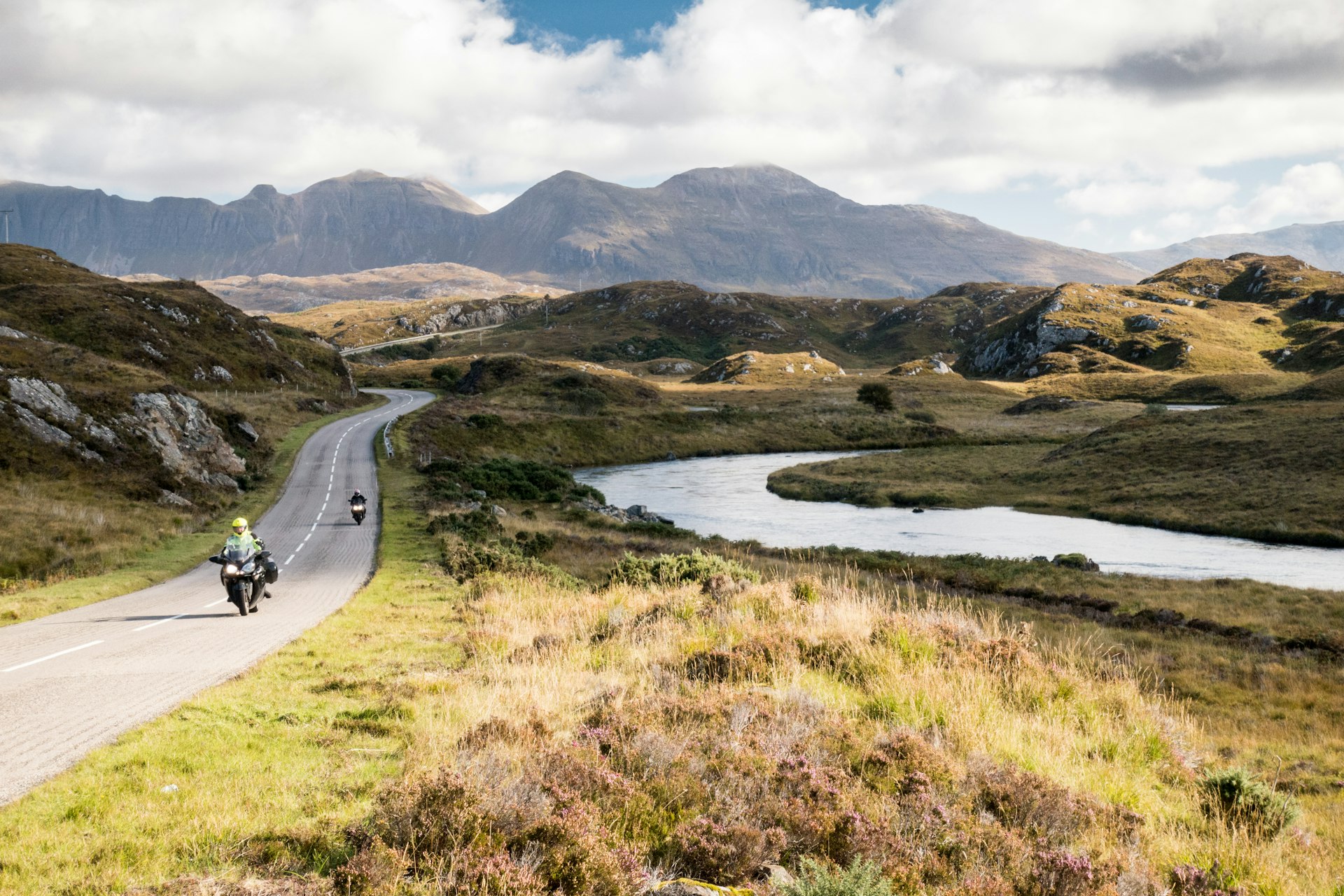 Motorcyclists drive down a road through the lush Scottish Highlands. 