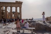 A tourist uses a hand fan to cool down another one sitting on a bench in front of the Parthenon at the ancient Acropolis in Athens,