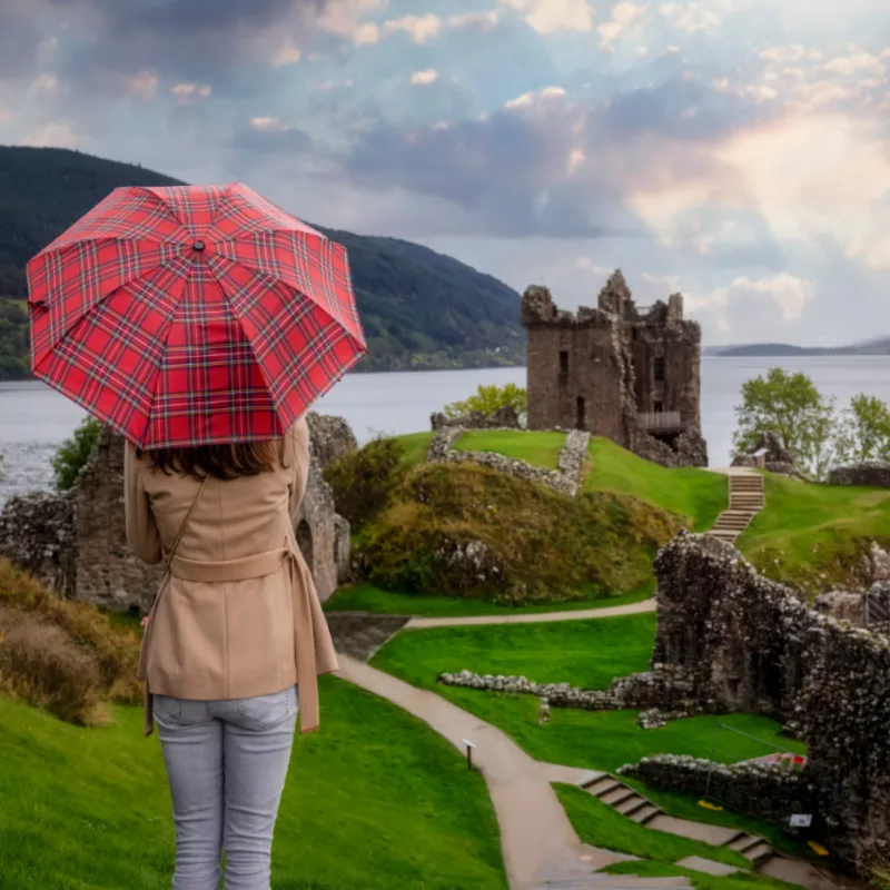 A tourist woman with a scottisch pattern umbrella looks at the famous Urquhart Castle at Loch Ness during autumn time, Scotland