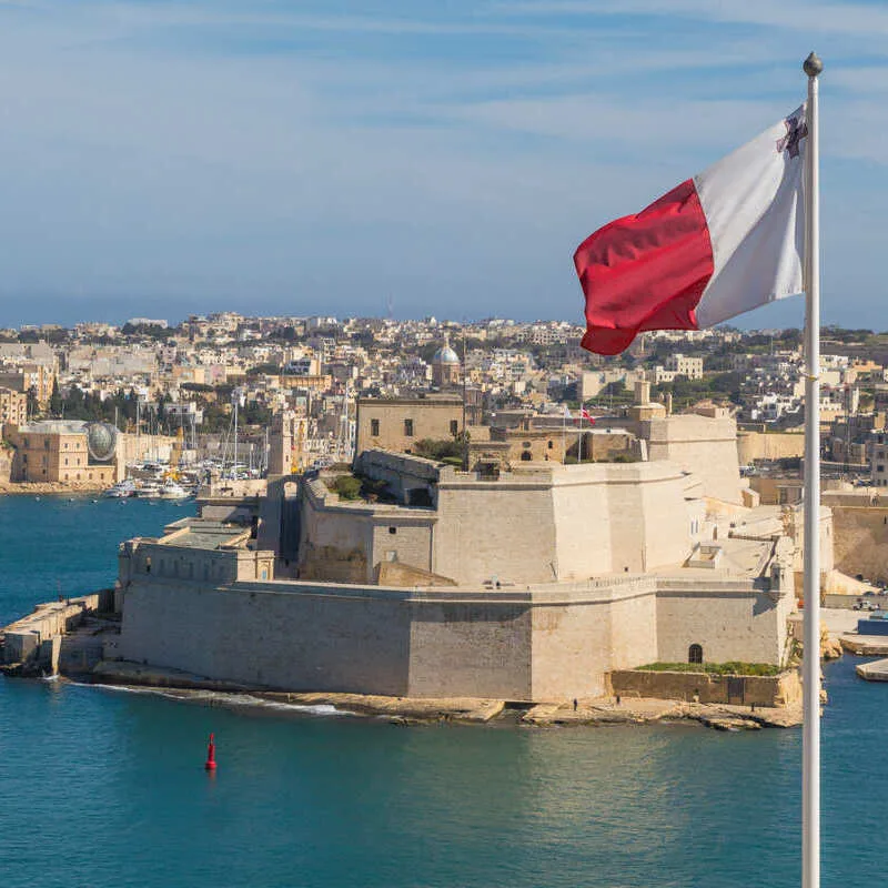 Maltese Flag Flying Against The Backdrop Of A Medieval City In Malta, Mediterranean Sea, Southern Europe