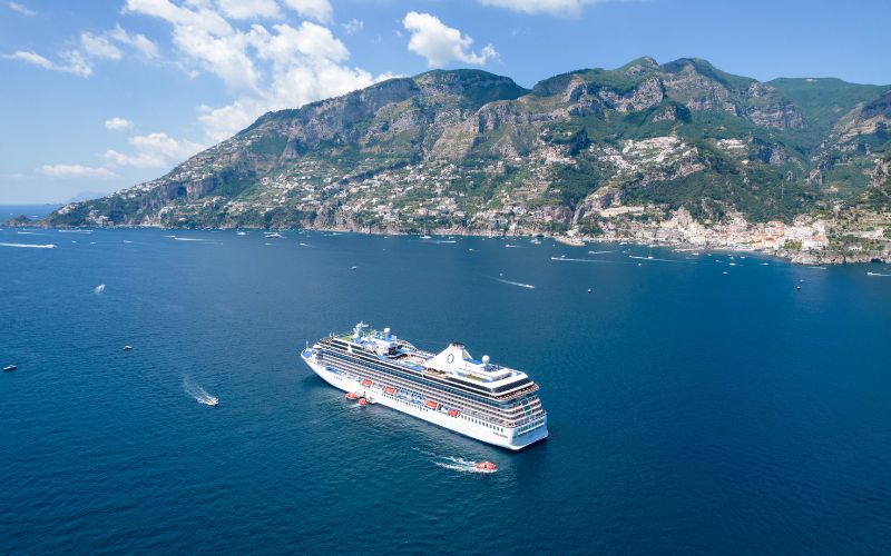 Oceania Riviera sails along the picturesque Amalfi Coast, with rugged mountains covered in greenery and quaint coastal towns visible in the background. The deep blue sea is dotted with small boats under a clear sky, adding to the serene and scenic landscape.