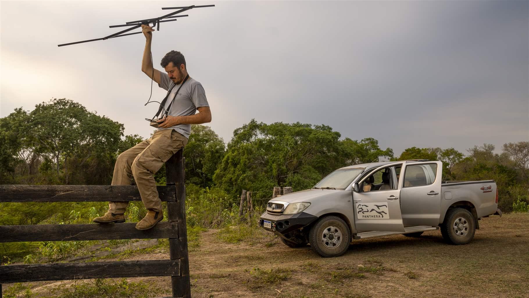 Under cloudy skies, Fernando Tortato sits astride a wooden fence in a small clearing, holding a small antenna aloft and looking down at a hand-held device. A few feet away, a silver pickup truck with both the front and back doors open on the driver’s side is parked alongside lush green trees. 