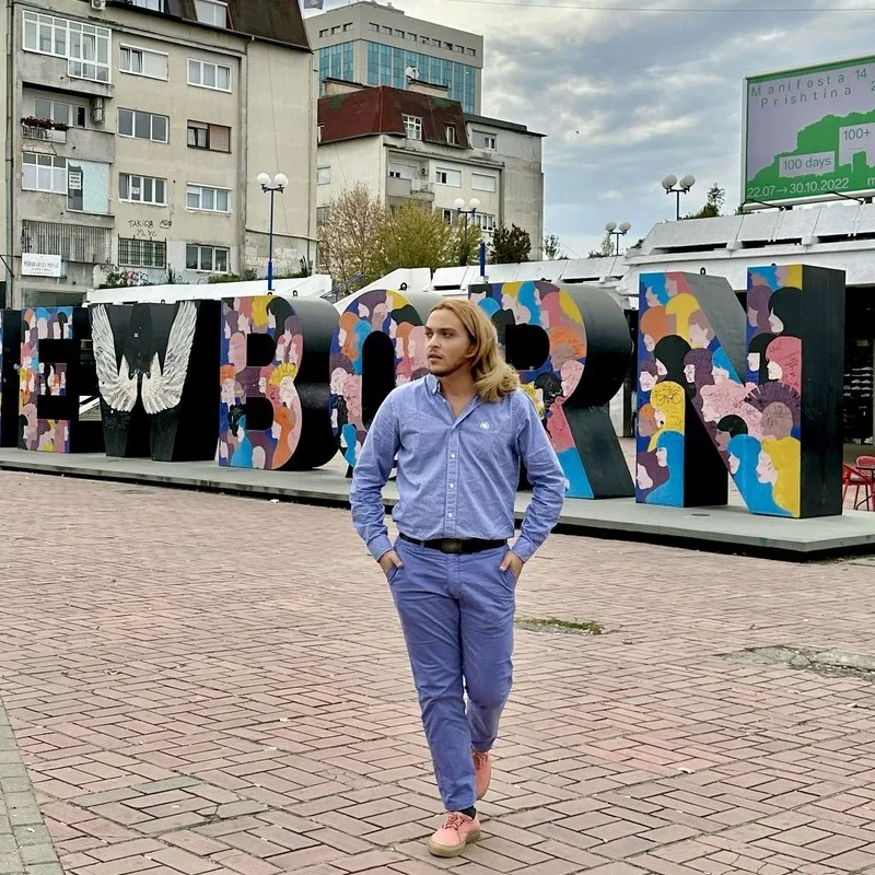 Young Male Tourist Posing For A Picture In Front Of The Newborn Sign In Pristina, The Capital City Of Kosovo, Eastern Europe