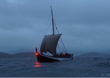 A recreation of a medieval era Norse ship sails on dark waters, with mountains in the background.