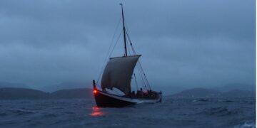 A recreation of a medieval era Norse ship sails on dark waters, with mountains in the background.